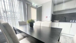 Ein moderner Essbereich mit einem dunklen Holztisch und weiß gepolsterten Stühlen, einer Zimmerpflanze sowie einer Küche mit weißen Schränken und einer schwarzen Arbeitsplatte.