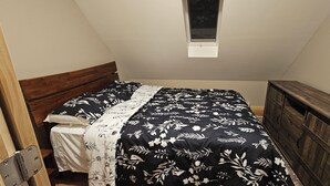 Une petite pièce triangulaire comprenant un lit, une tête de lit en bois, une petite fenêtre et une commode en bois.
