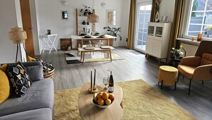 Una sala de estar moderna con un sofá gris, una mesa de centro de madera con fruta, una zona de comedor con una mesa blanca y vistas al exterior a través de grandes ventanas.