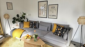 Una sala de estar moderna con un sofá gris, cojines amarillos y estampados, una pequeña mesa de centro de madera con frutas y flores, y dos cuadros enmarcados en la pared.