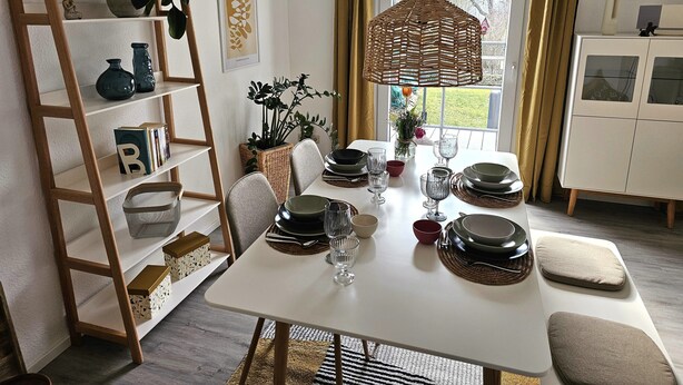 Un comedor con una mesa blanca dispuesta para cuatro personas, rodeado de una estantería de madera con objetos decorativos.