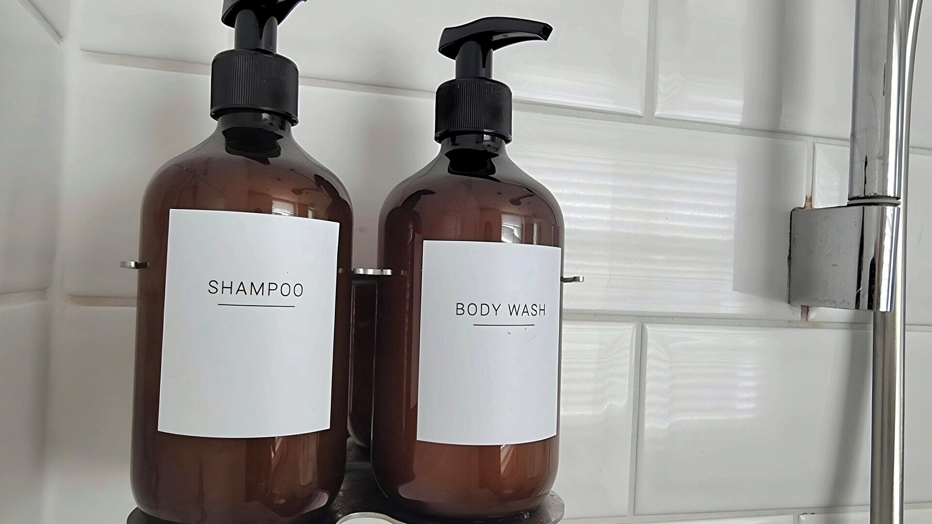 Dos botellas marrones con etiquetas blancas, una que dice "SHAMPOO" y la otra "BODY WASH", sobre una estantería en un baño con azulejos blancos.