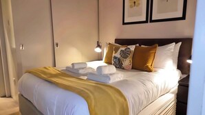 A neatly made bed with white linens and a yellow throw blanket, a bedside lamp, and a nightstand with a lamp. - *NEW* Notting Hill - It's The One One One! (2) (London)