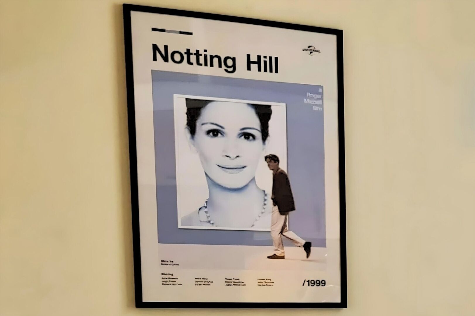A framed poster of Notting Hill with a blurred face, a person walking beside it, and text listing actors and the year 1999.
