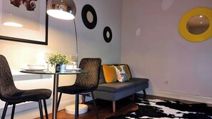 A modern living room with a glass dining table, two chairs, a grey sofa with colorful cushions, and a black and white cowhide rug.