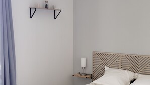 A neatly made bed with white linens and a tray of folded towels. There is a bedside table with a lamp and a small shelf above it. The room has a modern design with a geometric patterned headboard.