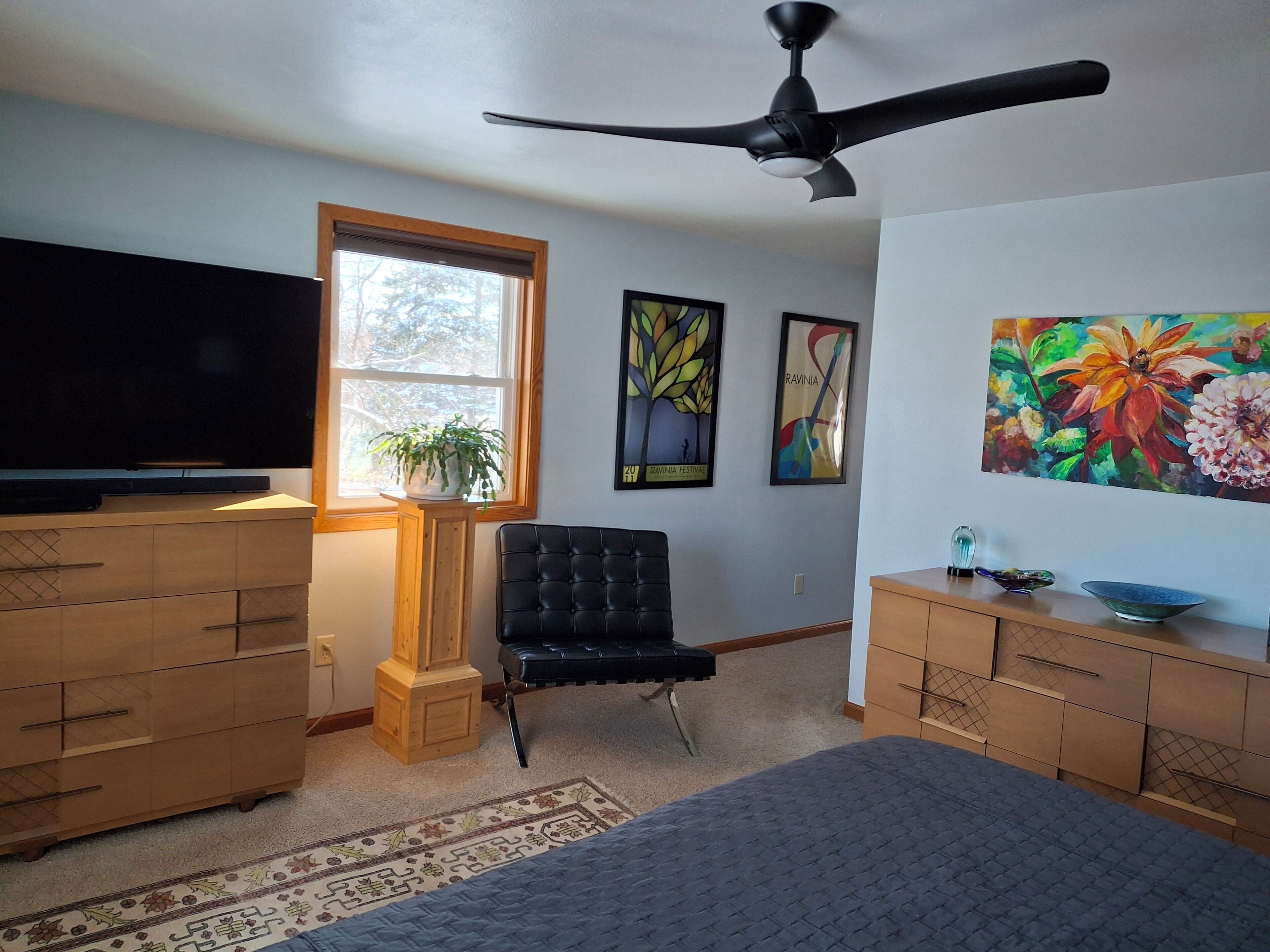 Een slaapkamer met een bed, een televisie, een plafondventilator, een raam met een plant en kunst aan de muren.