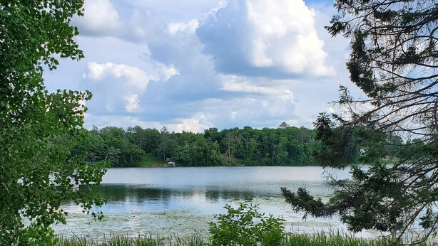 A wooden deck with two chairs and a table, overlooking a calm lake with dense greenery and trees in the background. - Private cabin with resort amenities; kayak, paddleboard, pontoon, great swimming (Pillager)