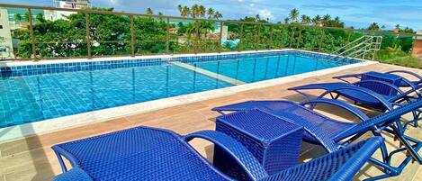 A rooftop pool with clear blue water, surrounded by blue wicker lounge chairs and a view of palm trees and buildings in the background.