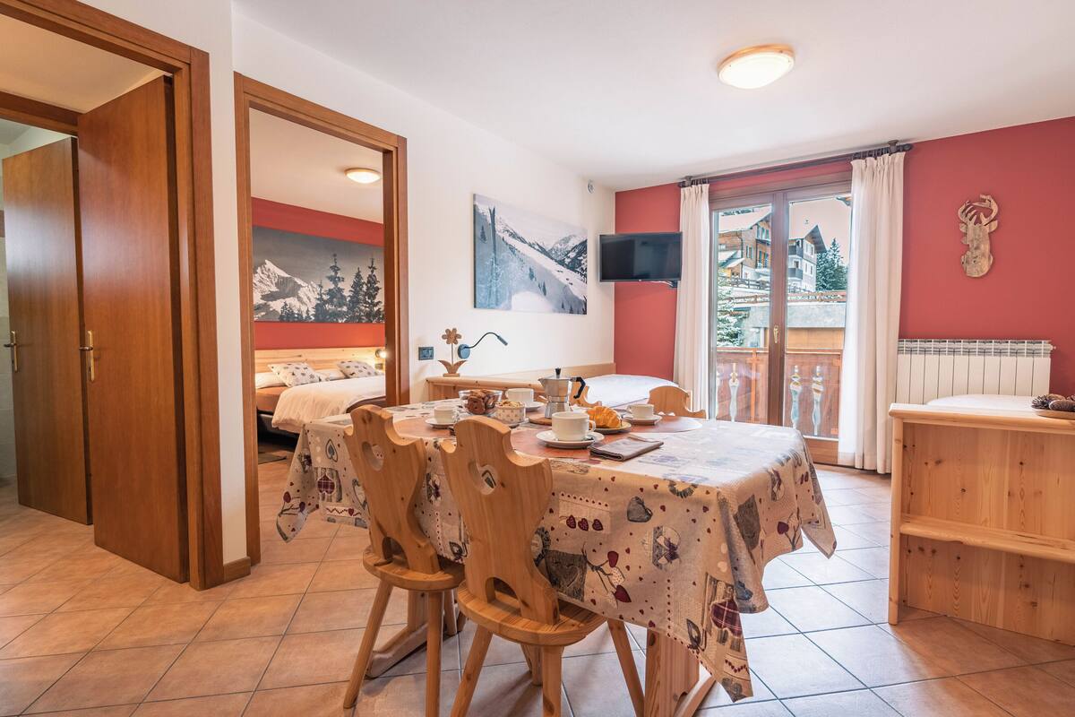 Un comedor con una mesa dispuesta para cuatro, sillas de madera, un cuadro de una montaña nevada y vistas a un edificio por una puerta de vidrio.