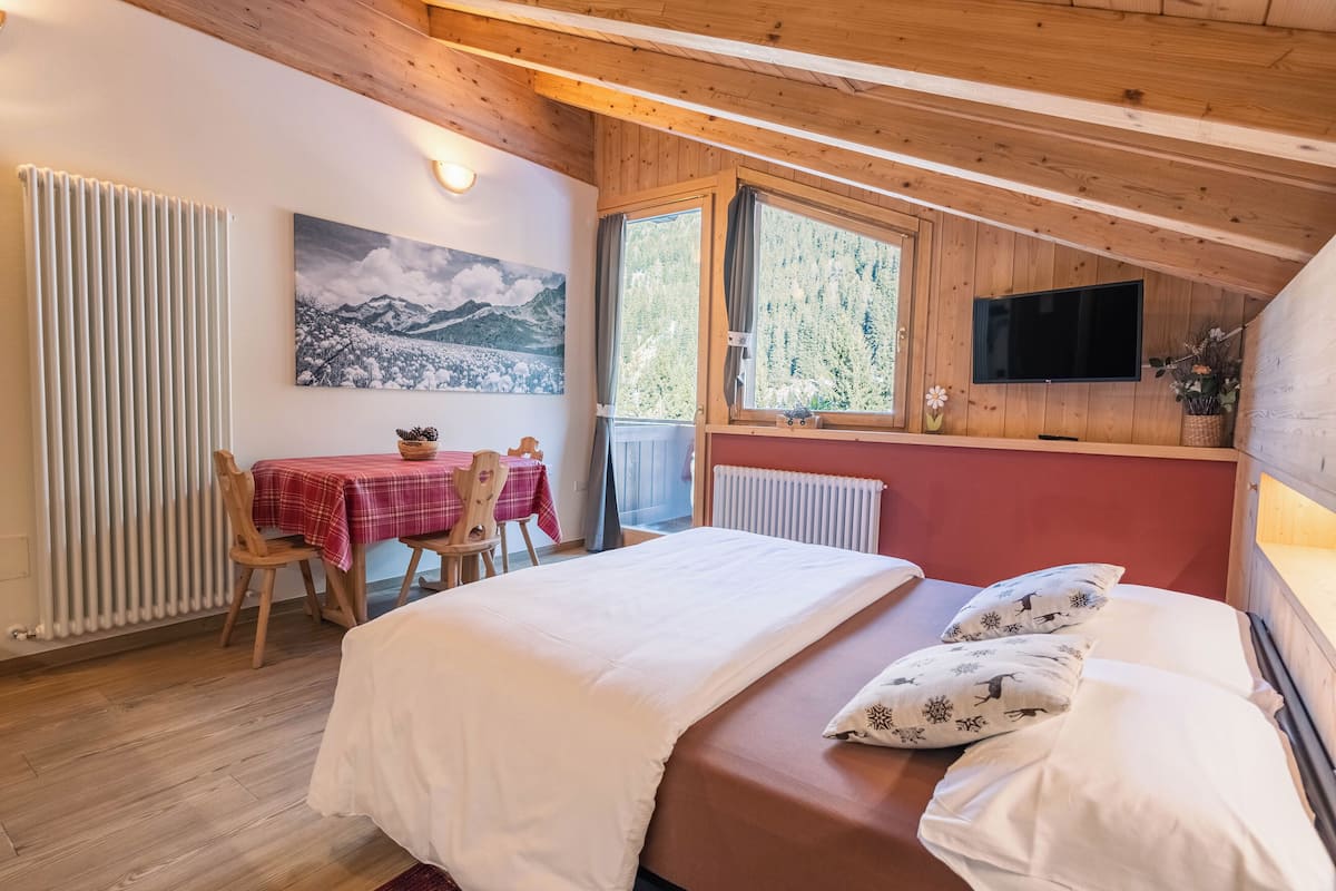 Una habitación acogedora con una cama, una mesa con sillas, un cuadro, un televisor y una ventana con vistas a árboles.