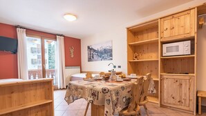 En hyggelig spiseplads med et træbord, stole og et buffetbord. Der er et træskab med en mikroovn og hylder. Et vindue med gardiner er synligt, og et maleri hænger på væggen.