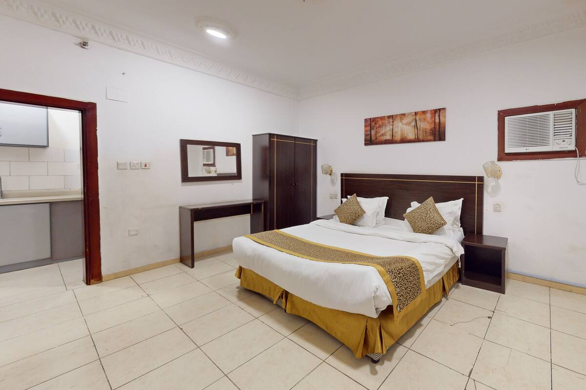 A neatly arranged bedroom with a large bed, a bedside table, a wardrobe, a mirror, and a painting on the wall.