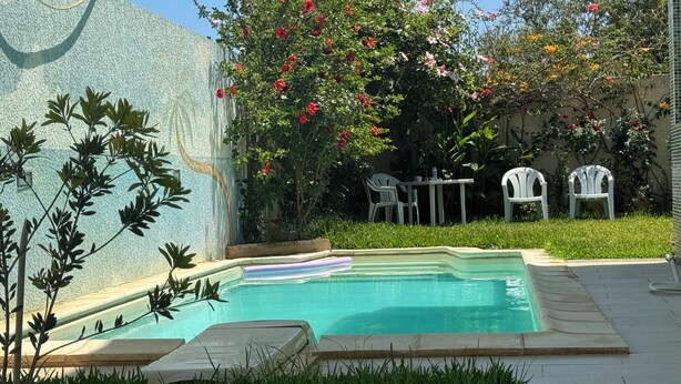 A swimming pool with a tiled deck, surrounded by grass and plants. There are white chairs and a table near the pool. - Villa 250 M2 on 800 M (Bizerte)
