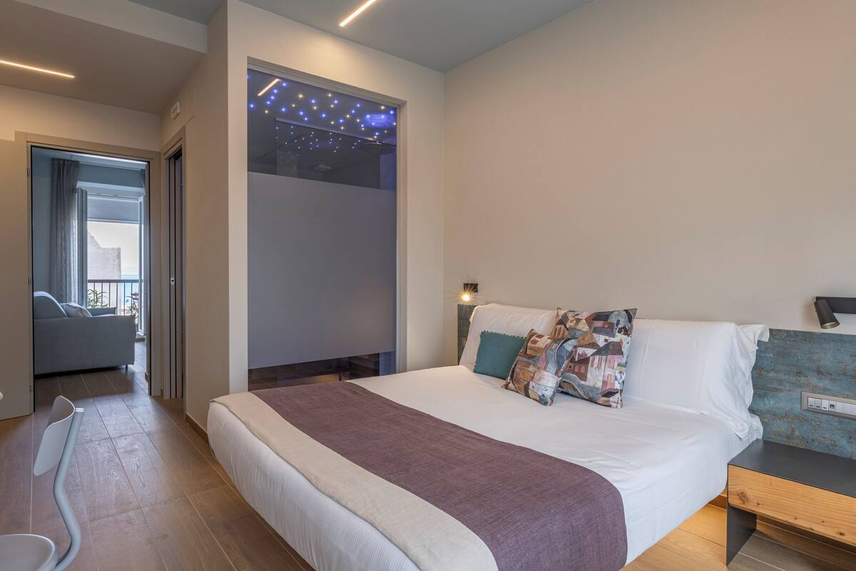 A modern hotel room with a large bed, a bedside table, a nightstand, and a view of the outdoors through a window.