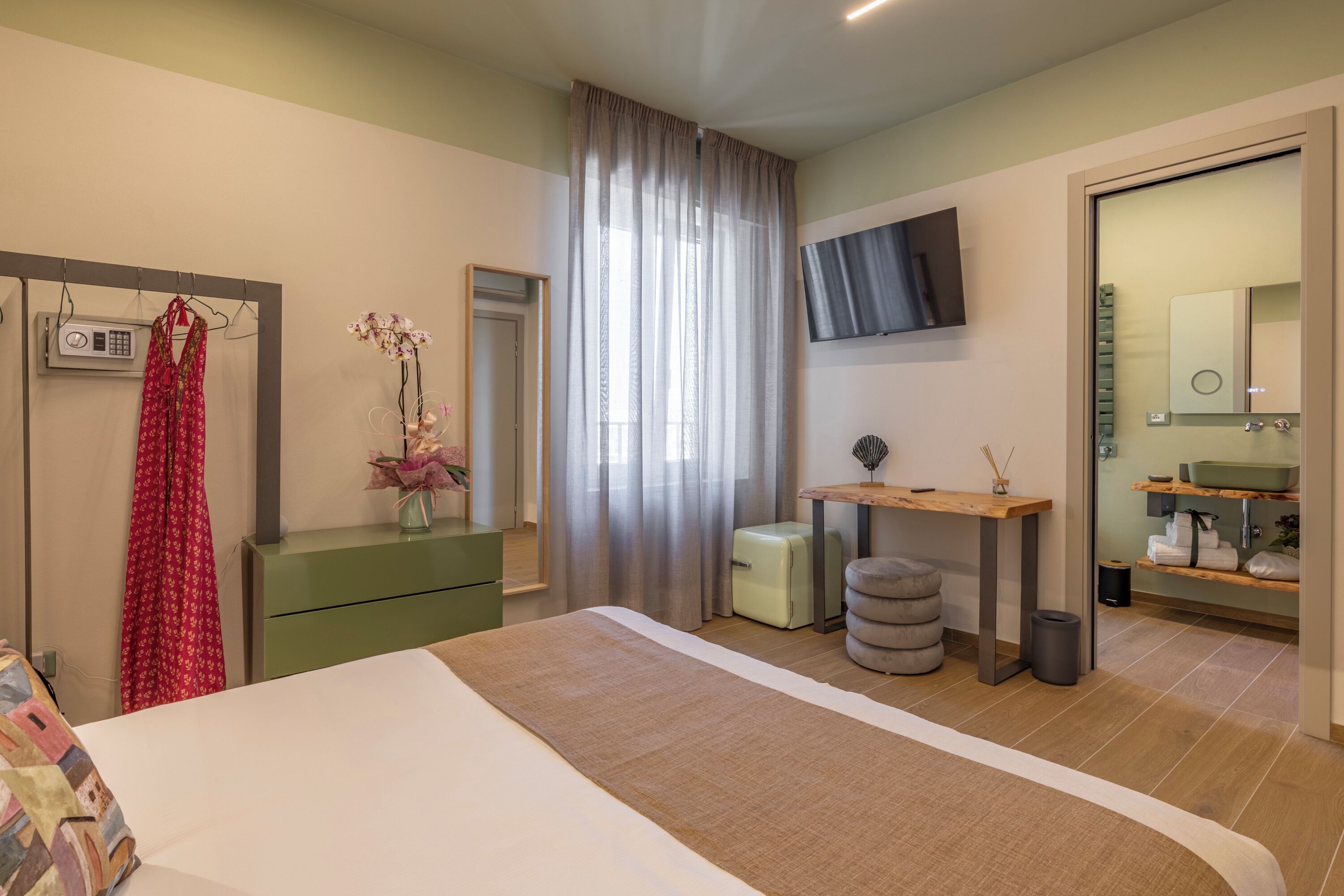 A modern bedroom with a large bed, a vanity with a mirror, a television mounted on the wall, and a small table with a vase of flowers.