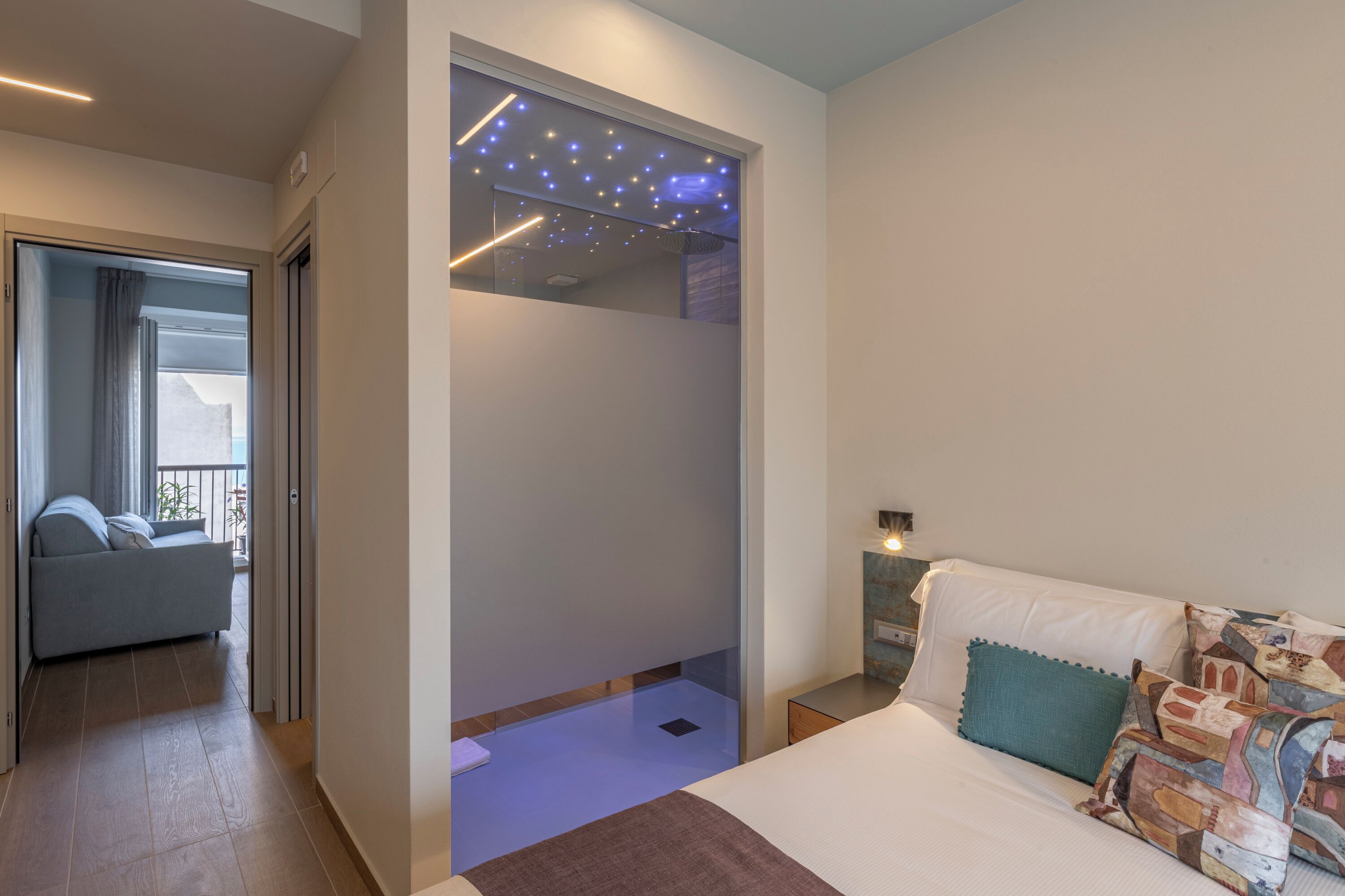 A modern bedroom with a bed, a nightstand, a sofa, and a balcony view.