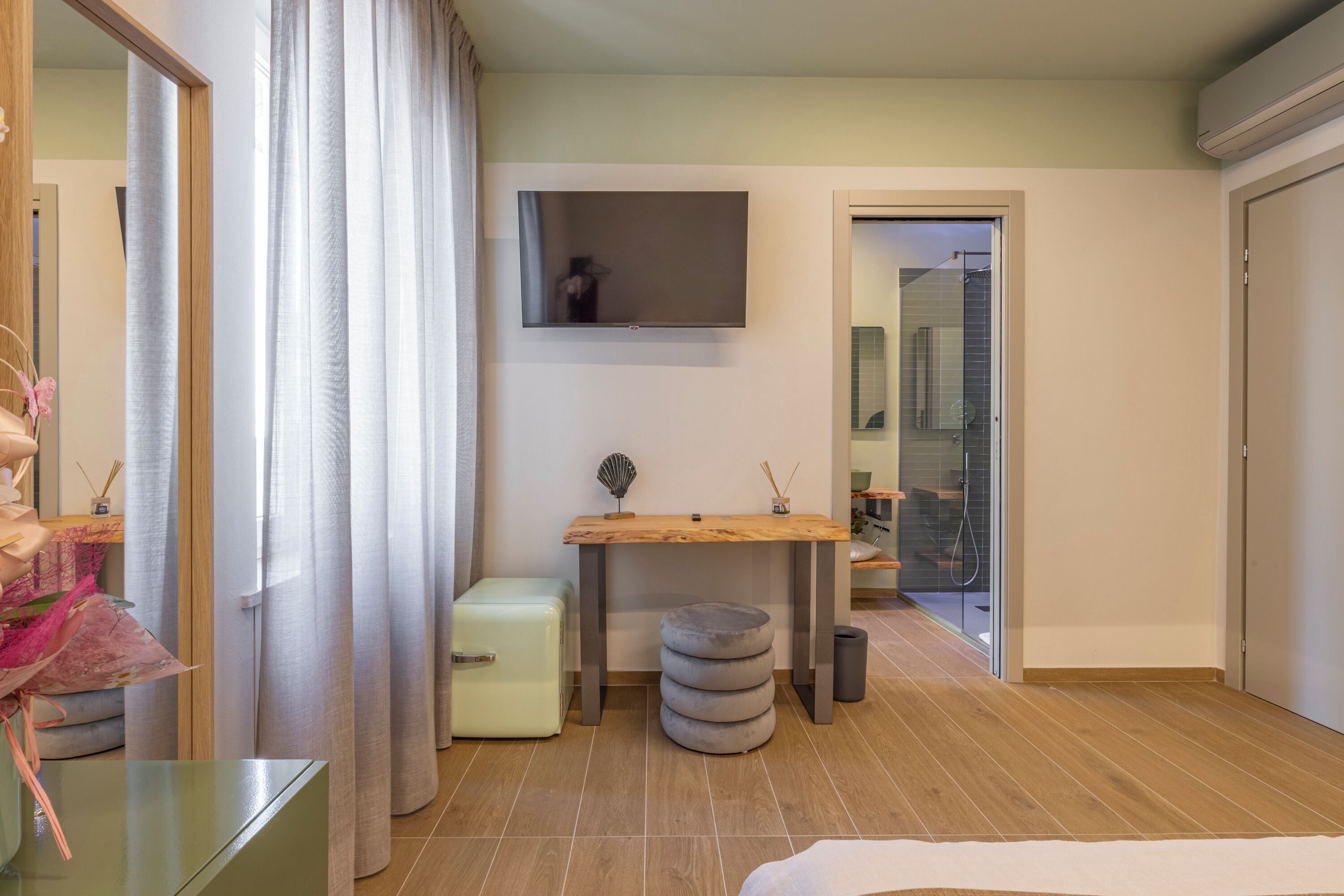 A modern hotel room with a wooden floor, a small table with a flat-screen TV, a compact fridge, and a large window with sheer curtains.