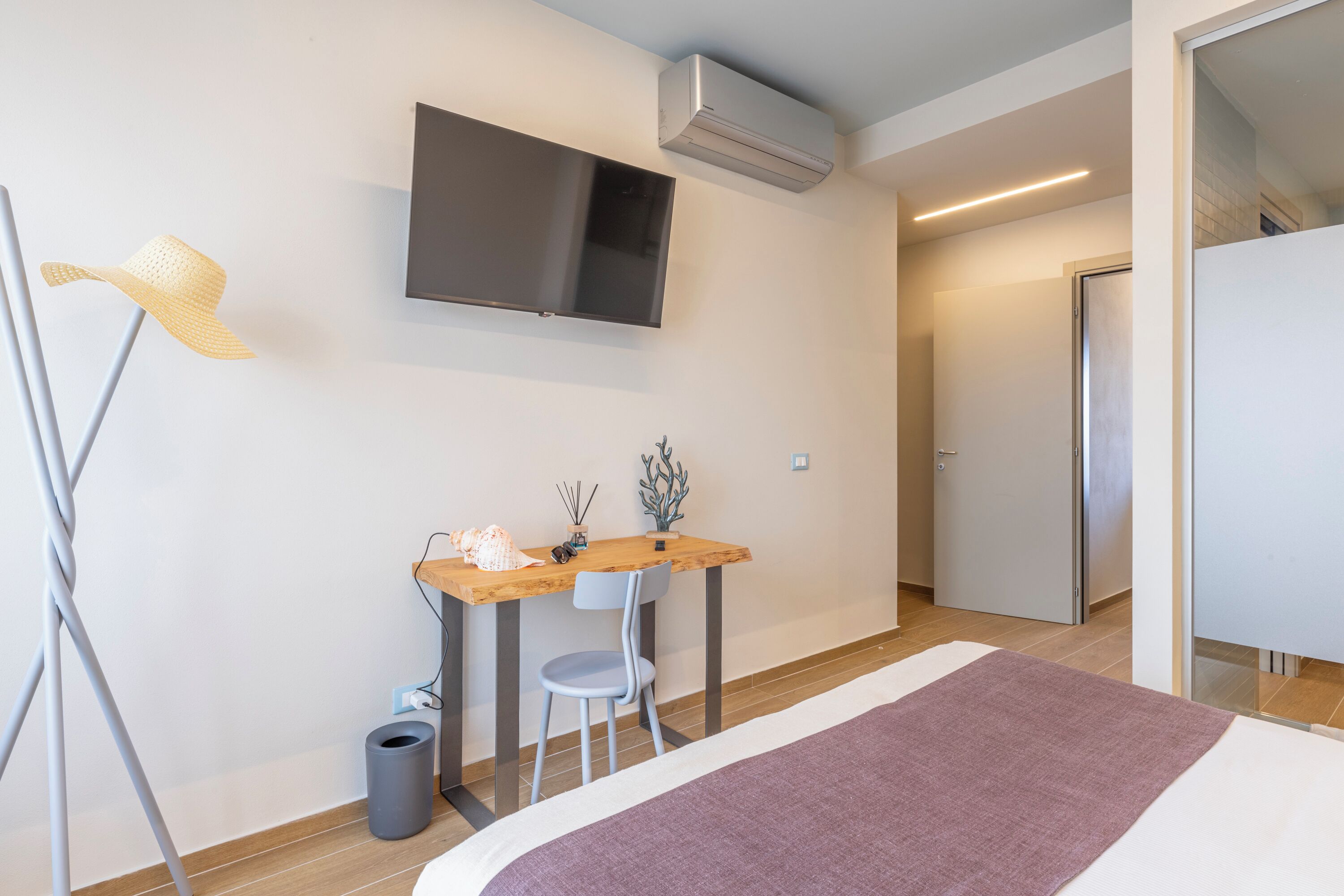 A modern hotel room with a bed, a desk with a chair, a television, and an air conditioning unit.