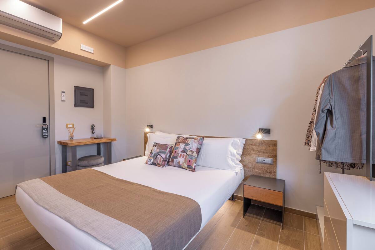 A modern hotel room with a large bed, bedside tables, a wardrobe, and a small desk with a chair.