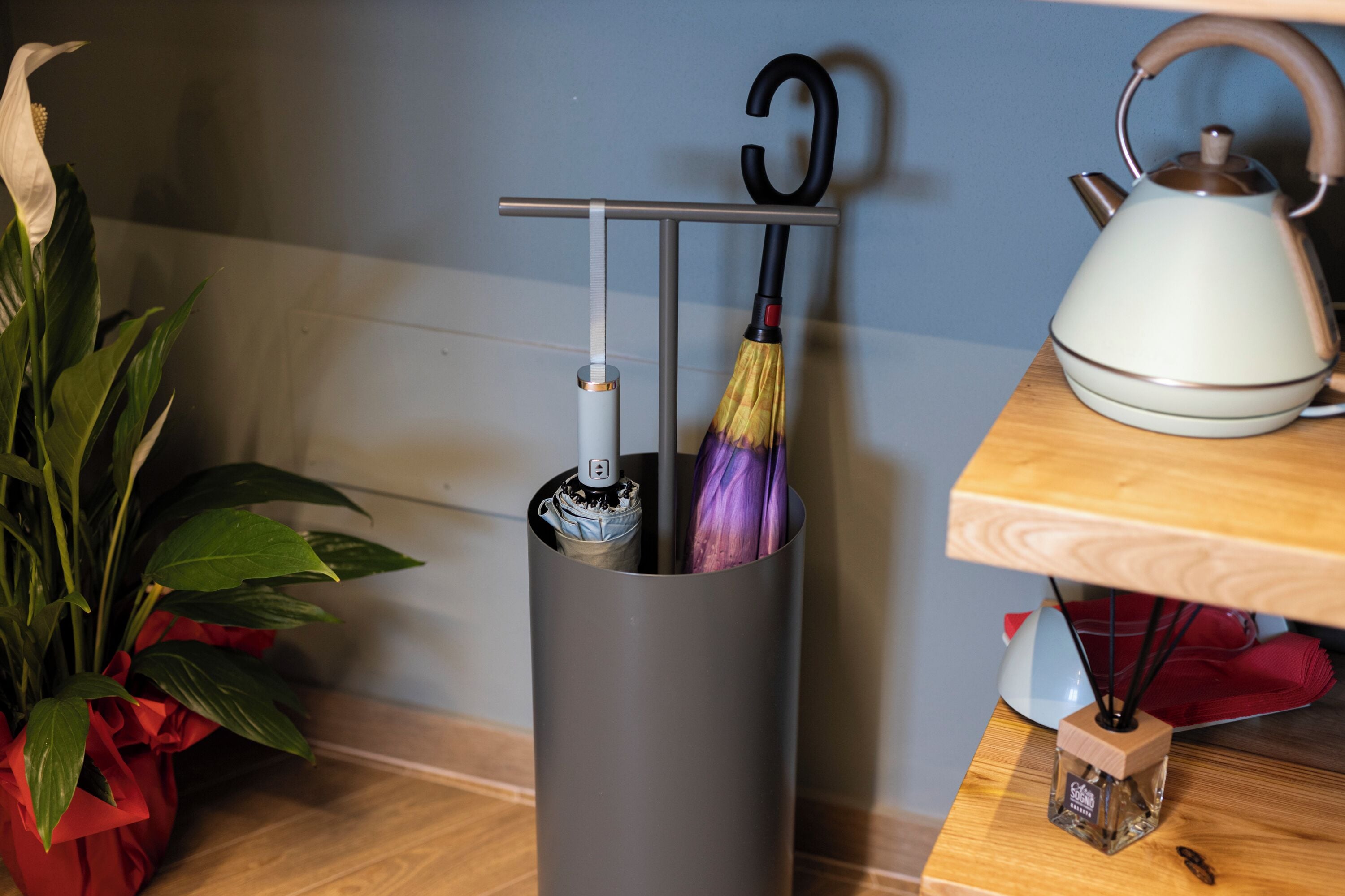 A grey vertical storage unit with a hook for hanging an umbrella, a white kettle on a wooden table, and a potted plant with red ribbons.
