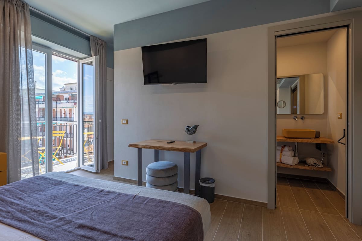 A modern hotel room with a bed, a TV mounted on the wall, a small wooden table, and a bathroom with a mirror and sink.