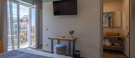 Ein modernes Hotelzimmer mit einem Bett, einem an der Wand montierten Fernseher, einem kleinen Holztisch und einem Badezimmer mit Spiegel und Waschbecken.