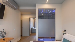 A modern hotel room with a bed, bedside table, and a small desk. There is a TV mounted on the wall, a window with curtains, and a door leading to another room. - Luci sul Mare (Follonica)