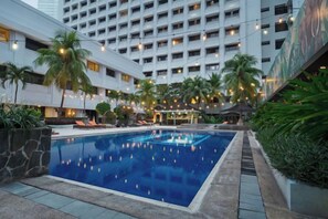 Un espace piscine d’hôtel avec une grande piscine, des palmiers et un bâtiment en arrière-plan.