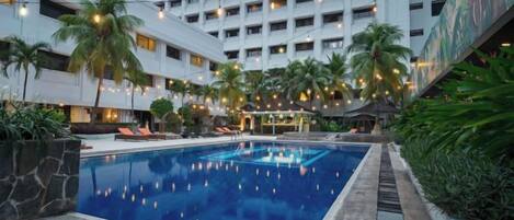 A swimming pool with a stone border, surrounded by palm trees and lounge chairs, with a modern building in the background.