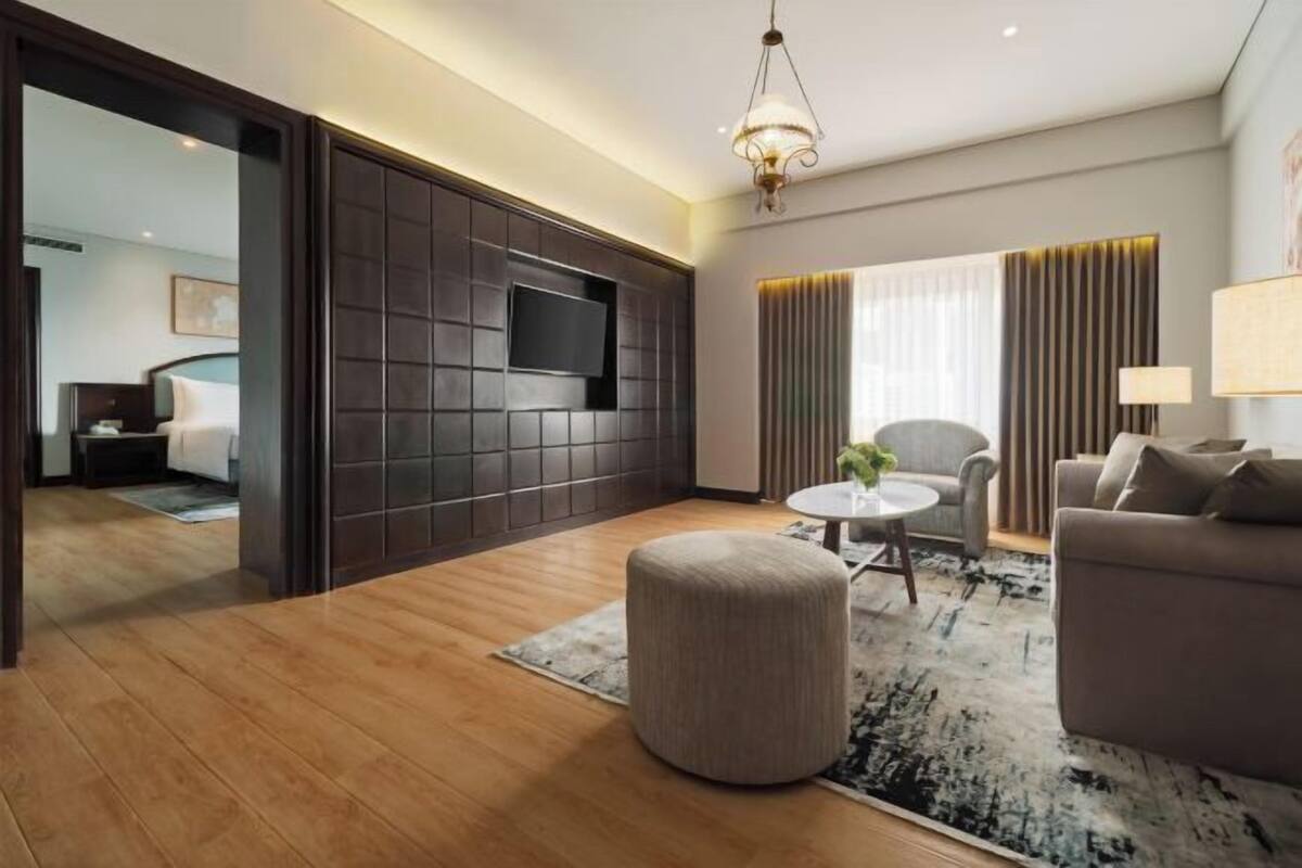 Een moderne hotelkamer met een houten vloer, een rond poefje, een bank, een televisie bevestigd aan een donkere paneelwand, een ronde tafel met een lamp, en uitzicht op een andere kamer met een bed.