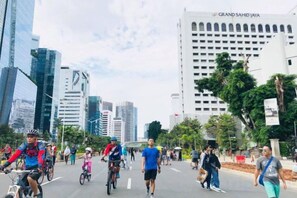 Une rue de la ville, avec des cyclistes et des piétons, des bâtiments modernes et, au fond, un hôtel imposant appelé « Grand Sahid Jaya ».