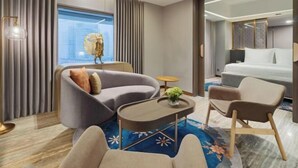 Ein modernes Hotelzimmer mit einer gebogenen Couch, einem kleinen runden Tisch, einem Bett und einem großen Fenster mit Vorhängen.