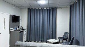 A modern hotel room with a grey sofa, a small table, a TV mounted on the wall, and a window with grey curtains. - Downtown inn  (Madaba)