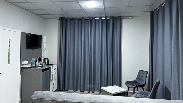 A modern hotel room with a grey sofa, a small table, a TV mounted on the wall, and a window with grey curtains. - Downtown inn  (Madaba)