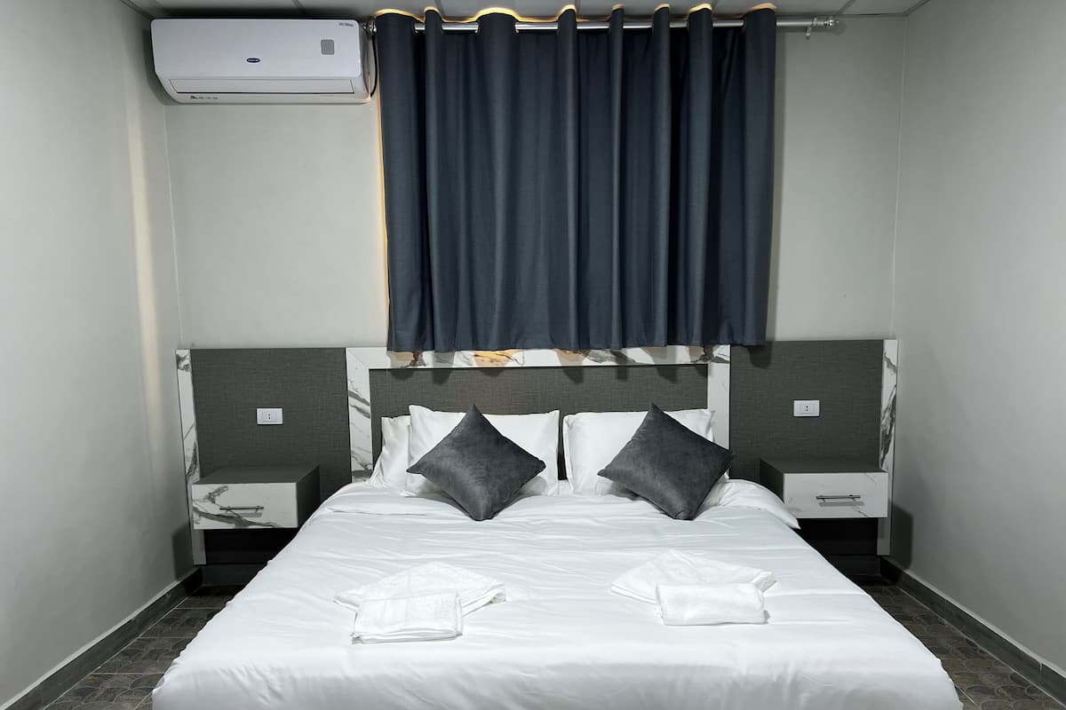 A neatly made bed with white linens and two pillows, a bedside table with a lamp, and a wall-mounted air conditioner.