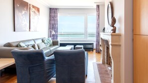 A modern living room with a sofa, two armchairs, a coffee table, and a fireplace. There is a large window with a view of the sea and a statue on the mantelpiece. - Apartment met Zeezicht in Oostende (Ostend)