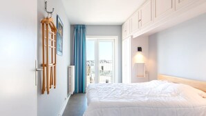 A bedroom with a large bed, white walls, a radiator, a window with curtains, and a view of buildings outside. - Apartment met Zeezicht in Oostende (Ostend)