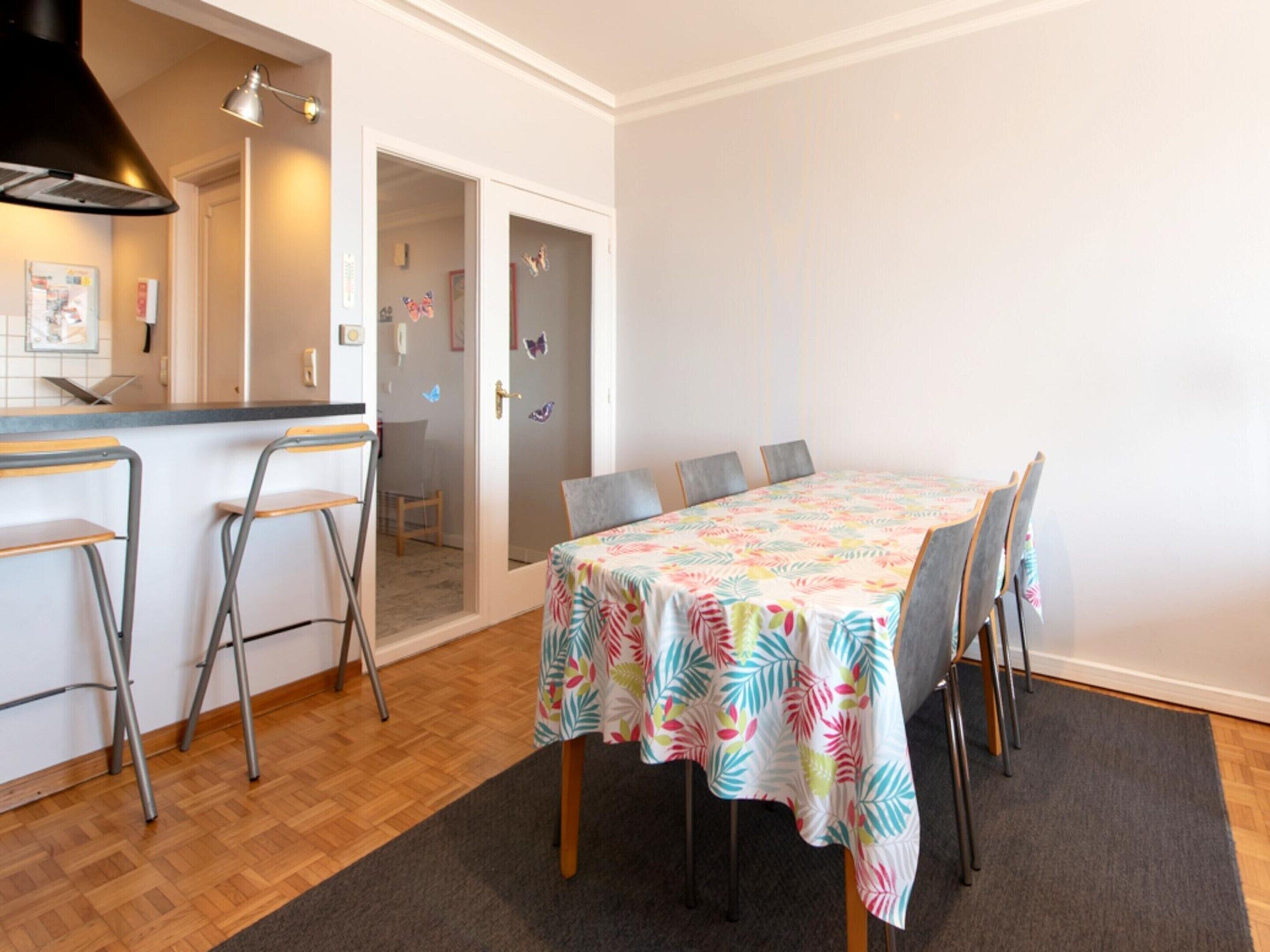 Un coin repas avec une table recouverte d’une nappe à motifs tropicaux, quatre chaises grises et un bar avec des tabourets.