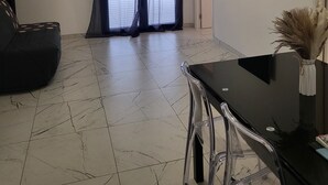 A modern living room with a black sofa, a dining table with chairs, a flat-screen TV mounted on the wall, and a large sliding glass door leading to an outdoor area.