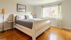 Een slaapkamer met een houten bed, een nachtkastje, een lamp, een lijst met een afbeelding aan de muur en een raam met gordijnen.