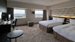 Habitación de hotel con dos camas, un escritorio, una silla y una mesita. Amplias ventanas ofrecen vistas a la ciudad.