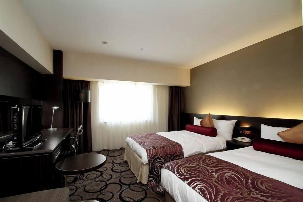 Ein Hotelzimmer mit zwei Betten, einem Schreibtisch mit Stuhl, einem kleinen Tisch und einem Fernseher.