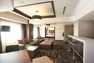 Habitación de hotel con sofá, mesa de comedor, zona de bar y dormitorio separado visible a través de una puerta abierta.
