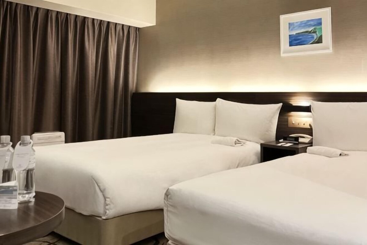 Habitación de hotel con dos camas, una mesita de noche con lámpara, un cuadro en la pared y vista al mar.