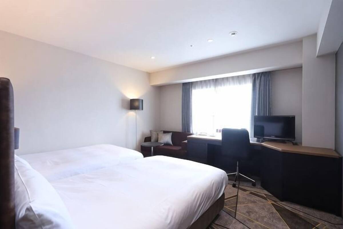 Ein Hotelzimmer mit einem großen Bett, einem Schreibtisch mit Stuhl, einem Fernseher und einem Fenster mit Vorhängen.