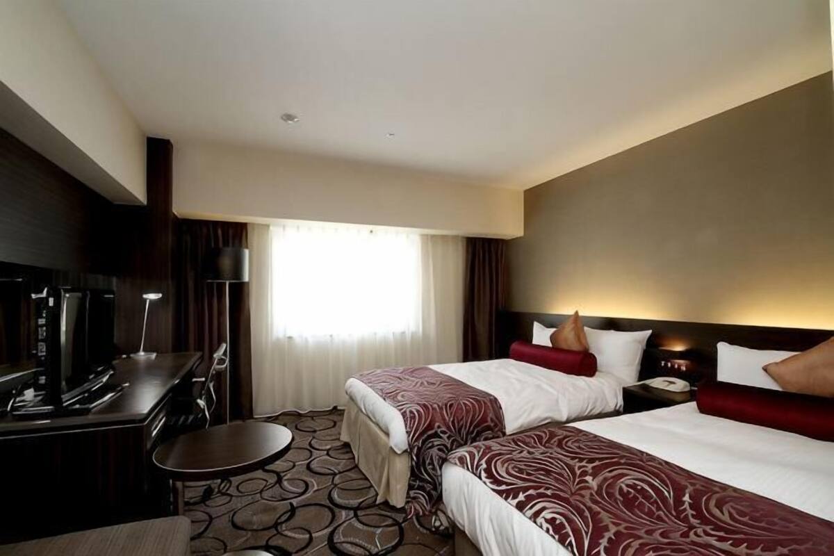 Ein Hotelzimmer mit zwei Betten, einem Schreibtisch, einem Stuhl, einem Fernseher und einem Fenster mit Vorhängen.