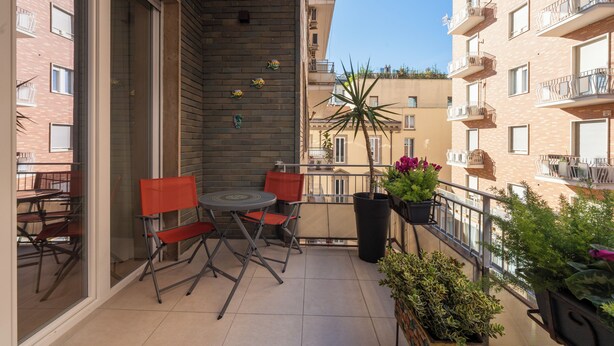 Uma varanda com mesa e cadeiras, plantas em vasos e vista para edifícios vizinhos.