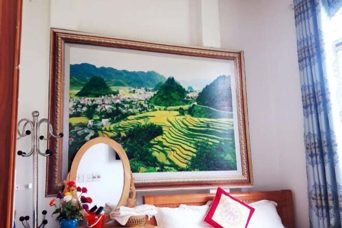 Un cuadro con un paisaje, una cama de madera, un espejo y un florero con flores sobre una mesita de noche.