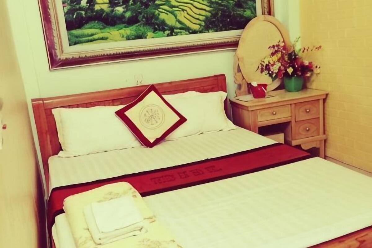 Una cama bien hecha con una manta roja, una mesita de noche de madera con un espejo y un jarrón de flores, y un cuadro en la pared.